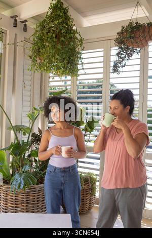 Mother and daughter standing on sunlit covered porch holding ceramic mugs next to lush plants Stock Photo
