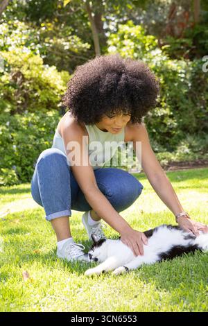 African american woman crouching on green lawn in private garden petting black-and-white cat Stock Photo