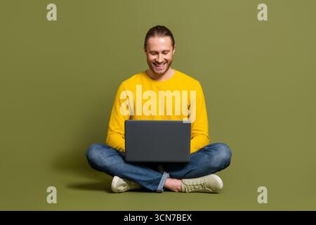Young man in casual yellow shirt smiling while using laptop sitting cross-legged against a minimalist green background Stock Photo