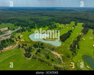 A high-angle aerial view of a beautifully maintained golf course with a large lake in the center, surrounded by a dense green forest. Stock Photo