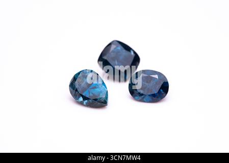 Three blue gemstones are sitting on a white background. The stones are all different shapes and sizes, but they all have a blue hue. The scene is calm Stock Photo