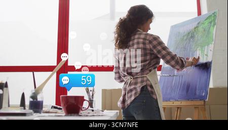 Artist wearing apron applying paint on blue green canvas in studio, with easel brushes, copy space Stock Photo