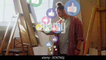 Standing middle-aged artist wearing coat in studio displaying canvas with floating thumbs-up icons Stock Photo