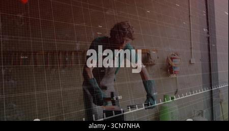 Sanding artisan wearing apron using power sander on wood plank in garage, digital grid overlay Stock Photo