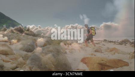 Traversing hiker carrying yellow backpack on boulders, sand near hillside with rainbow, copy space Stock Photo