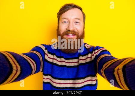 Cheerful bearded young man taking a selfie on a vibrant yellow background wearing a fashionable striped sweater Stock Photo