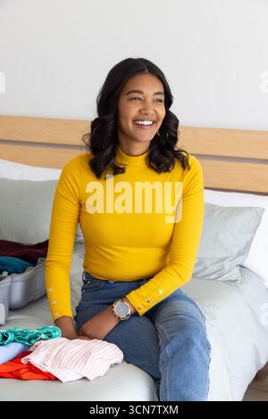 Woman sitting on bed smiling while packing suitcase in bedroom with folded clothes Stock Photo