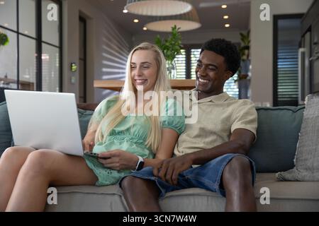 Diverse couple smiling and using laptop while sitting on sofa in modern living room Stock Photo
