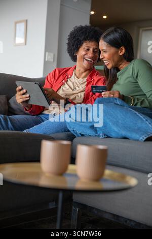 Diverse female friends leaning together on gray couch at home, browsing tablet, credit card Stock Photo
