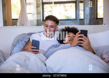 Diverse male partners reclining side by side on bed under duvet using smartphones in bedroom Stock Photo