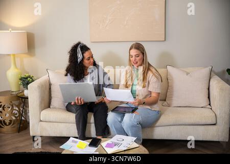 Diverse female colleagues collaborating over printed charts with tablet, laptop on sofa at home Stock Photo