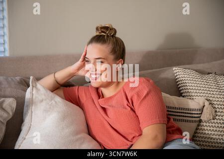 Woman reclining on sofa in living room with decorative pillows wearing coral top, denim jeans Stock Photo