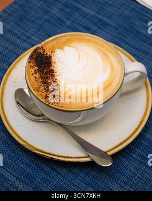 freshly made cappuccino on a textured blue placemat. The coffee is topped with a thick, frothy layer of foam, elegantly dusted with cocoa powder Stock Photo