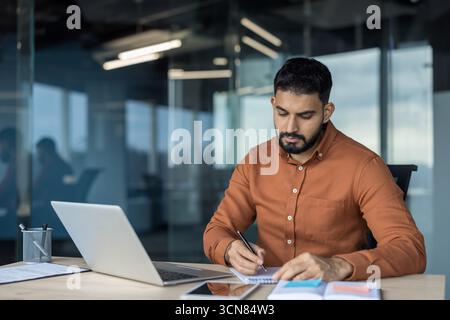 Professional businessman writing important notes in a notebook, concentrating on his work at a modern office desk with a laptop, tablet, and documents Stock Photo