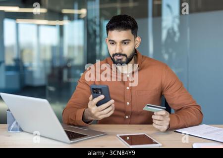 Young businessman making a secure online payment with smartphone and credit card at a modern office desk, engaged in ecommerce transaction using mobile banking app and wireless connectivity Stock Photo