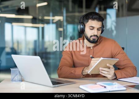 Young man with headphones works on a laptop in a modern glass-walled office, focused on taking notes in a notebook while studying or attending an online course for professional growth Stock Photo