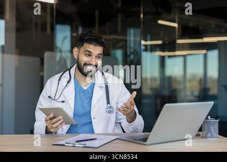 Male doctor in a lab coat with stethoscope conducts a telehealth video consultation from an office, using laptop and tablet to discuss patient care and medical advice remotely Stock Photo