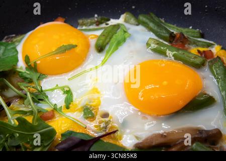 Close up of two fried eggs with bright yellow yolks served with green beans fresh herbs and vegetables in a black pan perfect for breakfast healthy ea Stock Photo