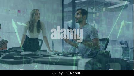 Standing man in gray polo holding tablet, gesturing in conference room with laptop, phones, graphs Stock Photo