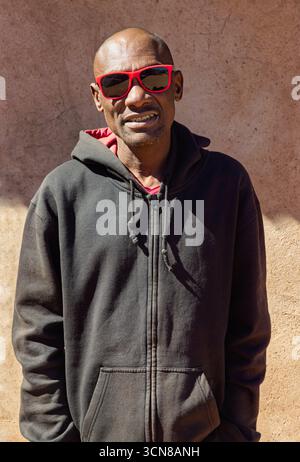 Africa village, single african young man, standing in front of the house in the yard, wearing red sunglasses Stock Photo