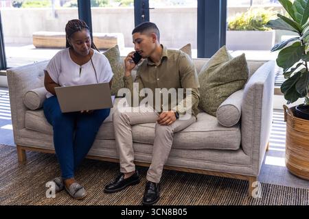 Diverse coworkers sitting on light gray sofa in office lounge using laptop and smartphone Stock Photo
