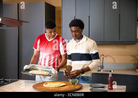 Diverse male friends preparing pizza in modern home kitchen using tray, oven mitt and fresh herbs Stock Photo