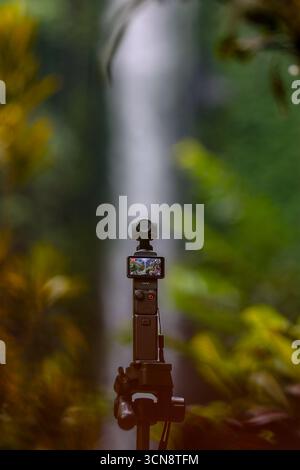 Gimbal camera mounted on a tripod with screen for video stabilization, capturing smooth footage of a waterfall scene in nature. Stock Photo