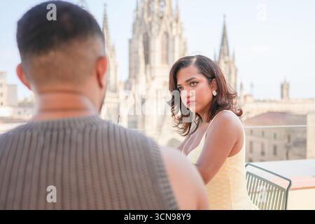 A man and a woman are sitting on a bench in front of a large building. The woman is wearing a yellow dress and has her hand on her hip. The man is loo Stock Photo