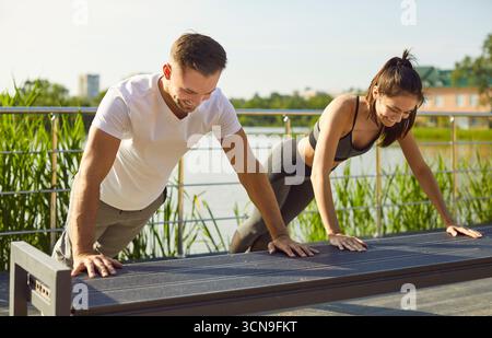 Smiling sporty young couple doing push ups on bench together outdoors, enjoying active healthy life. Stock Photo