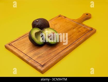 Close up of whole and sliced Avocados placed on a wooden board against a yellow background Stock Photo