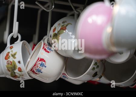 Vintage porcelain cups with floral and berry patterns hang on white hooks in a rustic setting. Made in GDR Stock Photo
