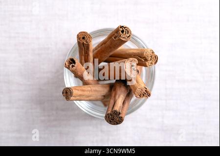 Dried Ceylon cinnamon bark strips, in a glass, on linen. Whole, dried bark quills of Cinnamomum verum. Stock Photo