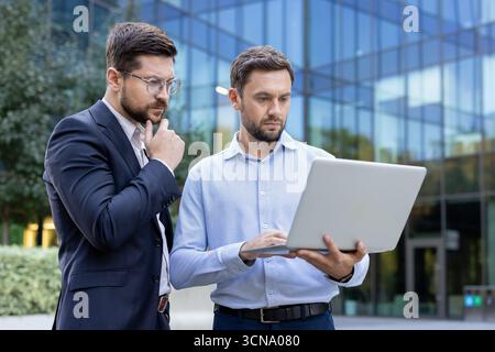 Two businessmen are collaborating and discussing ideas while one is holding and operating a laptop with the other thinking, both standing in front of a modern glass building Stock Photo