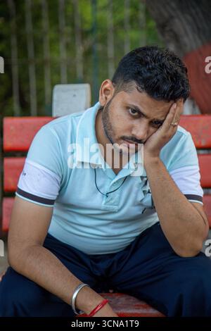 A man in a light blue polo sits on a red bench in a park, resting his head in his hand, conveying fatigue, worry, and a reflective mood. Stock Photo