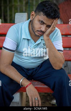A man in a light blue polo sits on a red bench in a park, resting his head in his hand, conveying fatigue, worry, and a reflective mood. Stock Photo