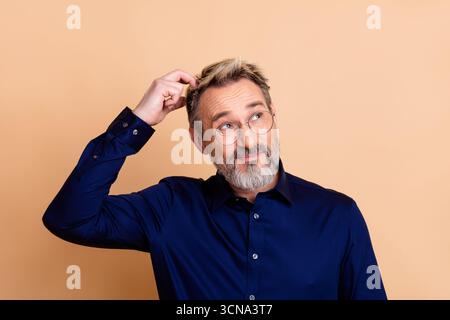 Portrait of a mature businessman in navy blue shirt scratching head with a puzzled expression against beige background Stock Photo