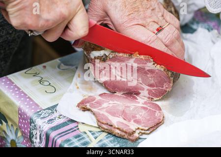 Hand cutting ham with knife. Fresh sliced meat on cutting board. Preparing food in kitchen. Cooking process and traditional meal preparation. Stock Photo