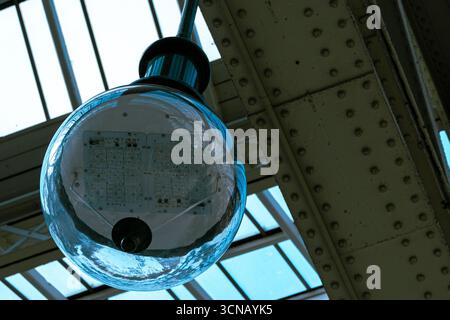 A close-up, low-angle shot of a modern LED globe light fixture hanging from an industrial ceiling structure with a glass roof. Stock Photo