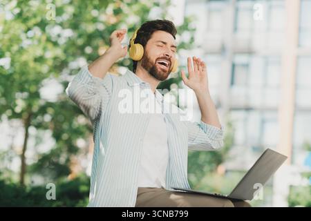 Cheerful young man enjoying music through headphones while working on a laptop outdoors in an urban setting Stock Photo