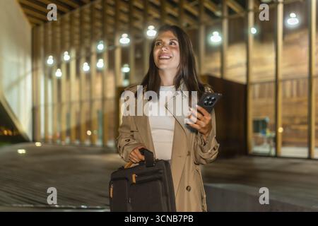 Young businesswoman walking confidently at night in an urban setting, carrying a briefcase and using a smartphone, embodying determination and modern Stock Photo