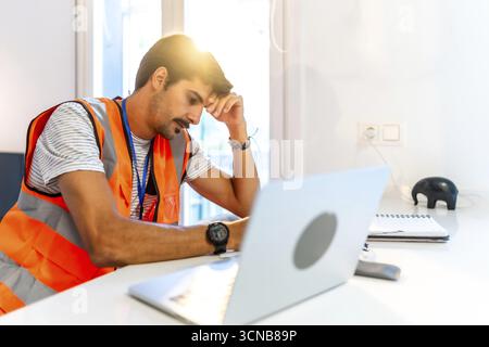 Young construction worker feeling stressed while working remotely from home using laptop, having problems and headache Stock Photo