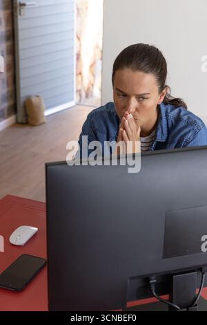 Woman sitting at home office desk wearing denim shirt focusing on monitor, mouse smartphone Stock Photo