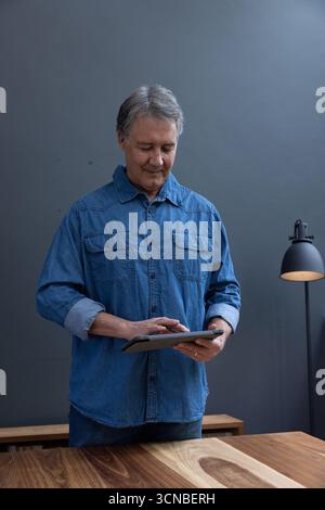 Senior man using tablet while standing behind wooden desk in room with lamps Stock Photo