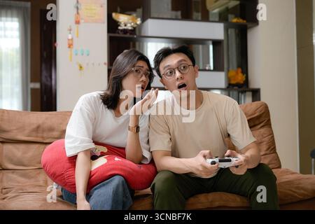 A couple sits close on a brown sofa, smiling while playing video games with handheld controllers Stock Photo