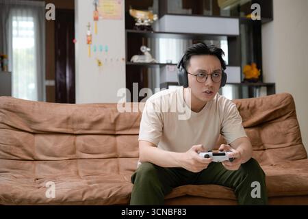 A young man sits on a brown couch wearing large headphones, gameplay on a gaming controller Stock Photo