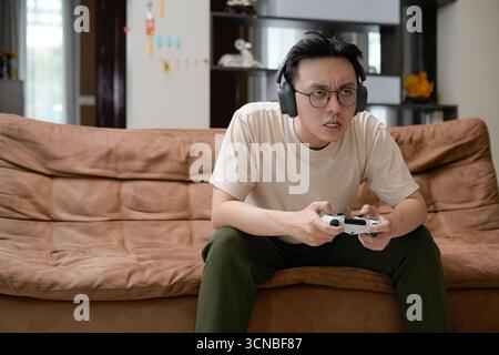 A young man sits on a brown couch wearing large headphones, gameplay on a gaming controller Stock Photo