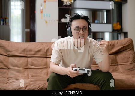A young man sits on a brown couch wearing large headphones, gameplay on a gaming controller Stock Photo