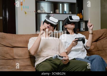 Two friends relax on a sofa at home, wearing VR headsets and using controllers to explore immersive virtual worlds Stock Photo