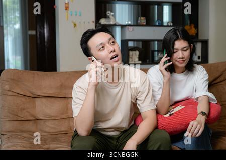 A young man and woman sit close on a sofa, talking on their smartphones in a cozy living room Stock Photo