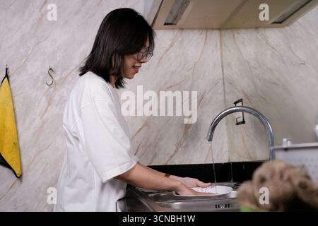 A young woman in a white tee washes dishes at a sleek kitchen sink Stock Photo